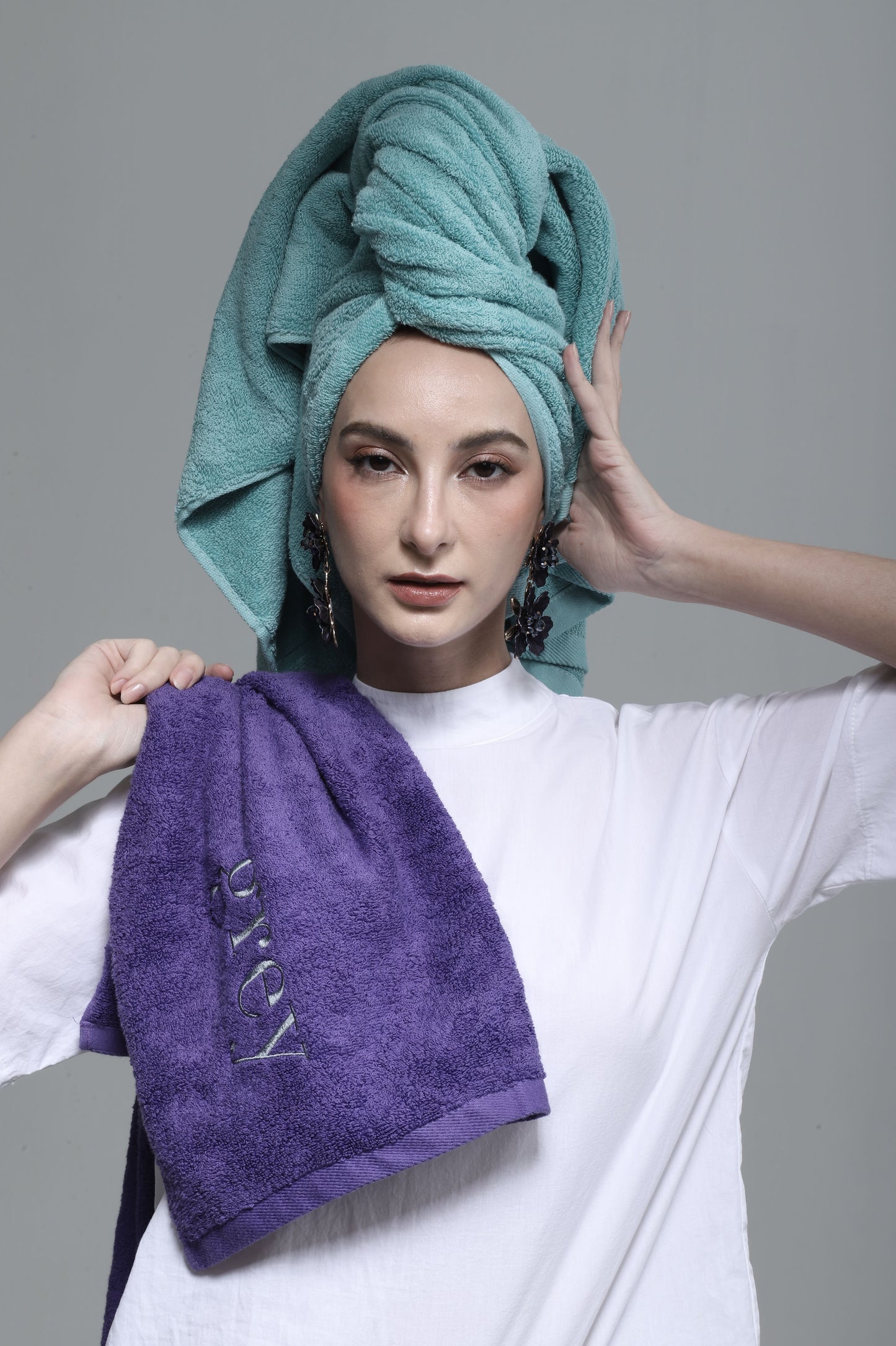 Person holding a purple towel with "GREY" branding, wearing a teal towel on their head against a gray background.
